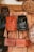 Several handcrafted leather bags and backpacks in various shades of brown and black are hanging on a wooden door. The bags feature intricate patterns and designs. Surrounding the bags are large decorative plates with carved geometric designs.