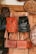 Several handcrafted leather bags and backpacks in various shades of brown and black are hanging on a wooden door. The bags feature intricate patterns and designs. Surrounding the bags are large decorative plates with carved geometric designs.