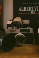 A vintage camera is prominently displayed on a wooden surface, surrounded by other camera equipment and accessories. In the background, a poster or book reads 'Albertville 1992' with some French text, suggesting an Olympic event theme. The setting conveys a nostalgic, analog atmosphere.