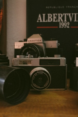 A vintage camera is prominently displayed on a wooden surface, surrounded by other camera equipment and accessories. In the background, a poster or book reads 'Albertville 1992' with some French text, suggesting an Olympic event theme. The setting conveys a nostalgic, analog atmosphere.