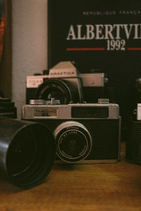 A vintage camera is prominently displayed on a wooden surface, surrounded by other camera equipment and accessories. In the background, a poster or book reads 'Albertville 1992' with some French text, suggesting an Olympic event theme. The setting conveys a nostalgic, analog atmosphere.
