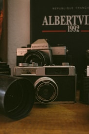 A vintage camera is prominently displayed on a wooden surface, surrounded by other camera equipment and accessories. In the background, a poster or book reads 'Albertville 1992' with some French text, suggesting an Olympic event theme. The setting conveys a nostalgic, analog atmosphere.