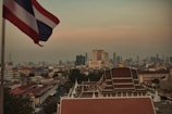 A cityscape featuring a skyline with modern skyscrapers in the distance and traditional Thai architecture in the foreground. A flag flutters prominently on the left side of the image, and the atmosphere appears hazy with a warm tone, possibly indicating dusk or dawn.