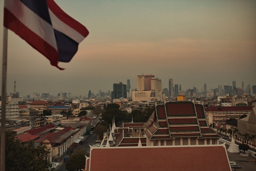 A cityscape featuring a skyline with modern skyscrapers in the distance and traditional Thai architecture in the foreground. A flag flutters prominently on the left side of the image, and the atmosphere appears hazy with a warm tone, possibly indicating dusk or dawn.