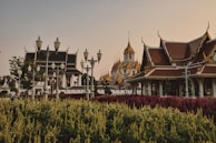 A scenic view of traditional Asian architecture with intricate rooftops and golden spires set against a backdrop of a clear sky. Lush gardens with vibrant red and yellow-green plants are visible in the foreground, while ornate street lamps line the pathway. People are casually walking along the path, enjoying the serene environment.