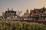 A scenic view of traditional Asian architecture with intricate rooftops and golden spires set against a backdrop of a clear sky. Lush gardens with vibrant red and yellow-green plants are visible in the foreground, while ornate street lamps line the pathway. People are casually walking along the path, enjoying the serene environment.