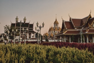 A scenic view of traditional Asian architecture with intricate rooftops and golden spires set against a backdrop of a clear sky. Lush gardens with vibrant red and yellow-green plants are visible in the foreground, while ornate street lamps line the pathway. People are casually walking along the path, enjoying the serene environment.