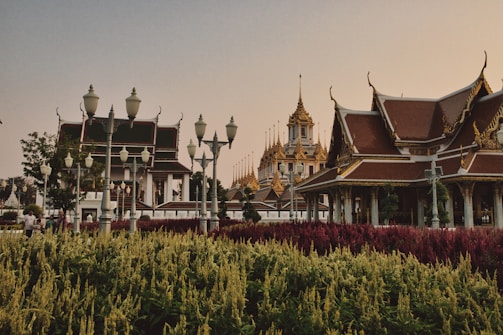 A scenic view of traditional Asian architecture with intricate rooftops and golden spires set against a backdrop of a clear sky. Lush gardens with vibrant red and yellow-green plants are visible in the foreground, while ornate street lamps line the pathway. People are casually walking along the path, enjoying the serene environment.