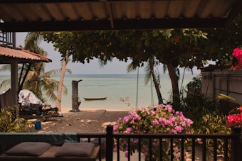 A serene beachfront view featuring a calm sea and a small boat anchored near the shore. Framed by lush green palm trees and vibrant pink flowers, the scene is set beneath a thatched roof. Soft sand leads to the tranquil water, while a few items like covered furniture and a blue container are visible in the shaded area of a patio.