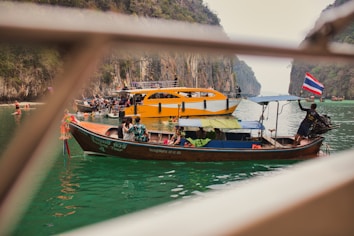 Multiple boats are seen navigating a tranquil turquoise waterbody surrounded by towering limestone cliffs. A longtail boat adorned with a Thai flag is positioned in the foreground, with several people onboard, one of whom appears to be steering. In the background, a larger orange and white boat is visible, carrying several passengers who appear to be tourists. The scene suggests a leisurely boating activity amidst a picturesque natural landscape.