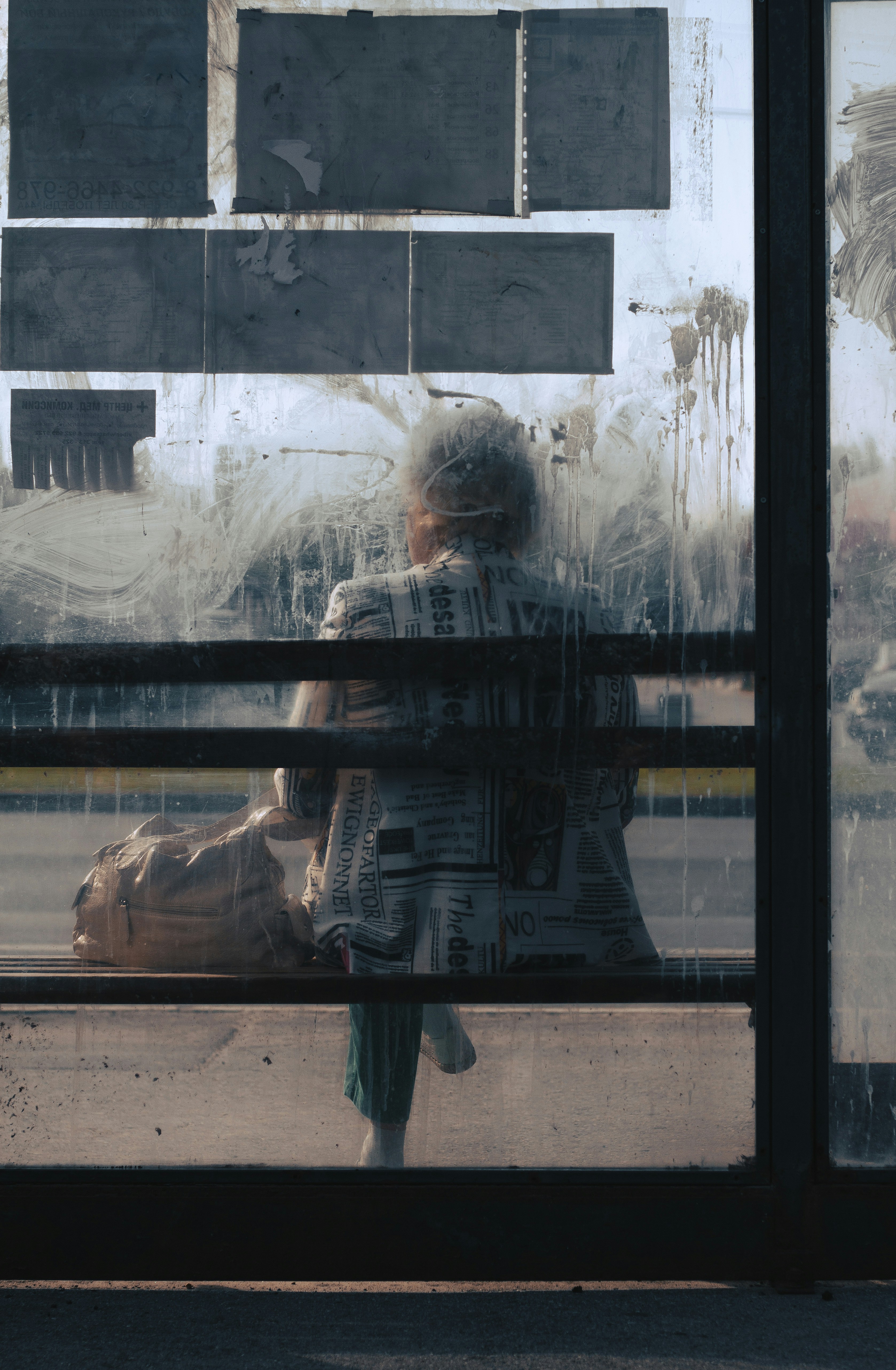 Woman reading a newspaper alone at a bus stop, pigeons around her feet, morning light filtering through trees