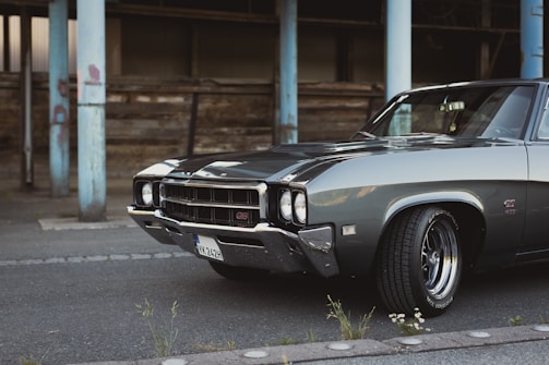 A classic muscle car parked on an asphalt road in an urban setting, showcasing a sleek, polished exterior. The car features chrome detailing, with a powerful front grille and prominent tires. In the background, there are weathered wooden structures and chipped blue metal pillars.