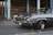 A classic muscle car parked on an asphalt road in an urban setting, showcasing a sleek, polished exterior. The car features chrome detailing, with a powerful front grille and prominent tires. In the background, there are weathered wooden structures and chipped blue metal pillars.