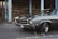 A classic muscle car parked on an asphalt road in an urban setting, showcasing a sleek, polished exterior. The car features chrome detailing, with a powerful front grille and prominent tires. In the background, there are weathered wooden structures and chipped blue metal pillars.