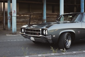 A classic muscle car parked on an asphalt road in an urban setting, showcasing a sleek, polished exterior. The car features chrome detailing, with a powerful front grille and prominent tires. In the background, there are weathered wooden structures and chipped blue metal pillars.