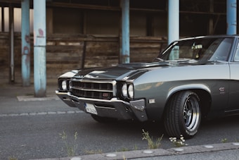A classic muscle car parked on an asphalt road in an urban setting, showcasing a sleek, polished exterior. The car features chrome detailing, with a powerful front grille and prominent tires. In the background, there are weathered wooden structures and chipped blue metal pillars.