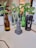 A collection of bottles and cans acts as makeshift plant pots for green leafy plants. The bottles vary in color, with green and brown glass, while one metal can has a prominent black color. In the foreground, a small gray sculpture resembling an Easter Island statue is placed. The background shows an indoor setting with blurred furniture and a person sitting at a table.