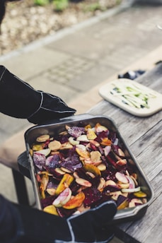 A person wearing dark gloves is holding a metal tray filled with a colorful mix of sliced vegetables, including beets, carrots, and other root vegetables. The tray rests on an outdoor wooden table, next to a cutting board with sliced cucumbers.