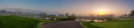 A serene landscape with a golf course under a misty morning sky. The rising sun casts a warm glow over the horizon, creating a gentle reflection on the water bodies. Lush green grass surrounds the area, and a golf cart is parked on the path.