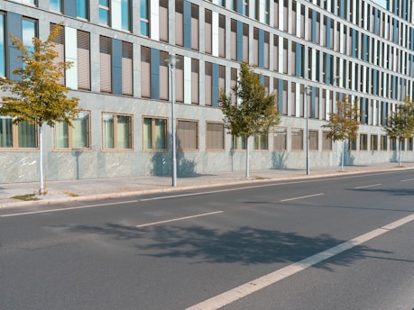 A modern office building with a grid of windows and a few small trees planted along the sidewalk. The street is empty with no cars or people visible.