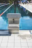 A long swimming pool with clear, blue water is lined with lane dividers in red and blue. At the forefront is a dive block numbered '1'. The background shows people, likely swimmers or pool-goers, gathered near the poolside under a sunny sky.