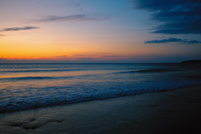 A serene beach at sunset, with gentle waves washing onto the shore. The sky is a gradient of warm orange and cool blue tones, with a few scattered clouds.