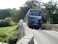A blue coach bus with travel agency branding is crossing a narrow stone bridge surrounded by lush greenery. In the distance, a few people can be seen walking on a country road.