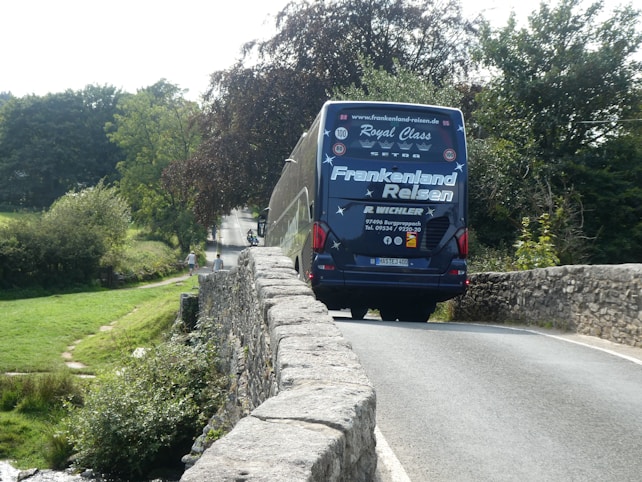 A blue coach bus with travel agency branding is crossing a narrow stone bridge surrounded by lush greenery. In the distance, a few people can be seen walking on a country road.