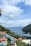 A scenic coastal view featuring a large yacht sailing on a calm blue sea. Luxurious red-orange umbrellas are spread around a pristine swimming pool overlooking the ocean. Lush green foliage surrounds the area, with a rocky hillside meeting the water. White clouds dot the bright blue sky.
