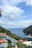 A scenic coastal view featuring a large yacht sailing on a calm blue sea. Luxurious red-orange umbrellas are spread around a pristine swimming pool overlooking the ocean. Lush green foliage surrounds the area, with a rocky hillside meeting the water. White clouds dot the bright blue sky.