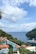 A scenic coastal view featuring a large yacht sailing on a calm blue sea. Luxurious red-orange umbrellas are spread around a pristine swimming pool overlooking the ocean. Lush green foliage surrounds the area, with a rocky hillside meeting the water. White clouds dot the bright blue sky.