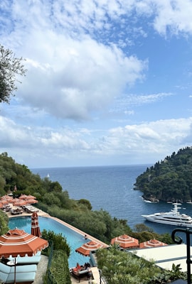 A scenic coastal view featuring a large yacht sailing on a calm blue sea. Luxurious red-orange umbrellas are spread around a pristine swimming pool overlooking the ocean. Lush green foliage surrounds the area, with a rocky hillside meeting the water. White clouds dot the bright blue sky.
