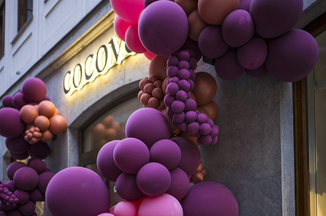 Clusters of vibrant balloons in shades of purple, pink, and brown are creatively arranged over a building entrance. A sign with illuminated letters spells out 'COCOV', partially obscured by the balloons. The background features a stone facade with a window.