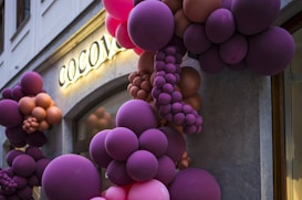 Clusters of vibrant balloons in shades of purple, pink, and brown are creatively arranged over a building entrance. A sign with illuminated letters spells out 'COCOV', partially obscured by the balloons. The background features a stone facade with a window.