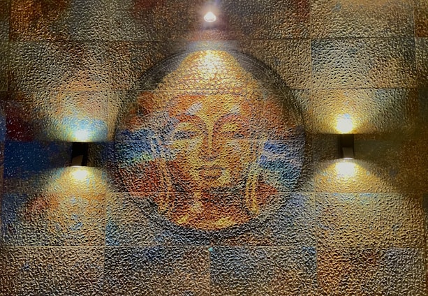 A textured wall art features a serene Buddha face in warm tones, illuminated by three soft lights. The artwork consists of a circular motif with detailed textures creating a mosaic effect.
