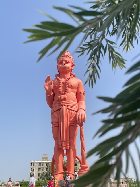 A large, orange statue of a mythological figure stands prominently against a clear blue sky. The statue is intricately detailed, featuring traditional clothing and holding a weapon. Framed by green foliage that appears in the foreground, the setting includes a few people walking nearby and a building in the background.