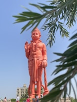 A large, orange statue of a mythological figure stands prominently against a clear blue sky. The statue is intricately detailed, featuring traditional clothing and holding a weapon. Framed by green foliage that appears in the foreground, the setting includes a few people walking nearby and a building in the background.