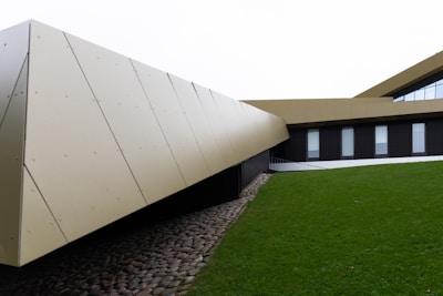 A modern architectural building features sleek, angular lines. The structure is clad in beige panels and integrated with dark vertical sections that incorporate windows. It sits on a neatly maintained green lawn, and a cobblestone path runs alongside the building, adding a touch of rustic contrast.