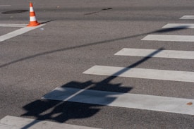 A traffic cone is positioned on an asphalt road next to a pedestrian crosswalk. The shadows of traffic lights stretch across the road surface, creating contrasting patterns on the pavement.