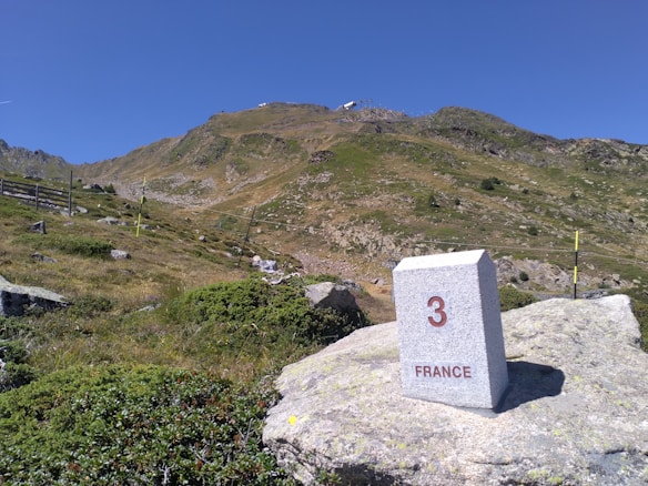 A picturesque mountain scene featuring a stone boundary marker with the number '3' and the word 'FRANCE' engraved on it. The foreground is covered in green vegetation and rocky terrain, leading up to a mountain slope with scattered rocks and patches of grass under a clear blue sky.
