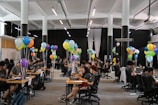 A large room filled with people sitting at desks working on laptops, accompanied by clusters of colorful balloons, under bright lighting in a spacious hall.