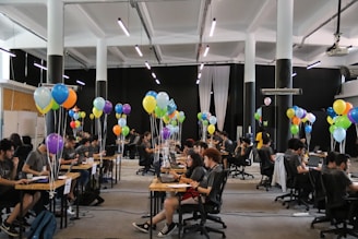 A large room filled with people sitting at desks working on laptops, accompanied by clusters of colorful balloons, under bright lighting in a spacious hall.