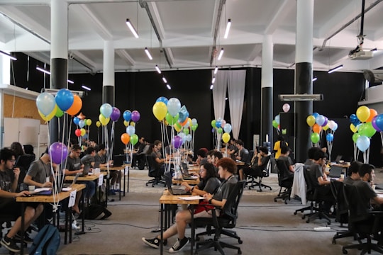 A large room filled with people sitting at desks working on laptops, accompanied by clusters of colorful balloons, under bright lighting in a spacious hall.