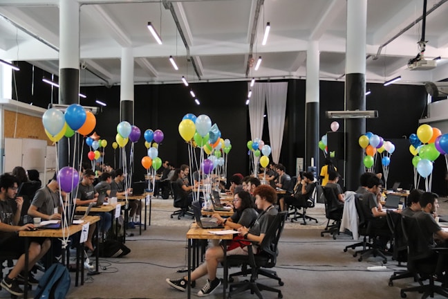 A large room filled with people sitting at desks working on laptops, accompanied by clusters of colorful balloons, under bright lighting in a spacious hall.