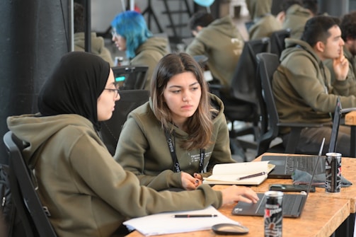 Two people are seated at a wooden table, working together on laptops. Several open notebooks and energy drink cans are visible. They appear to be in a collaborative office space with others working in the background.
