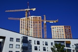 A construction site with two yellow cranes towering over newly constructed or under-construction high-rise buildings. These buildings have numerous windows and a modern design look. The foreground includes a multi-story building with unique geometric windows and a tree with green leaves.