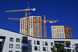 A construction site with two yellow cranes towering over newly constructed or under-construction high-rise buildings. These buildings have numerous windows and a modern design look. The foreground includes a multi-story building with unique geometric windows and a tree with green leaves.