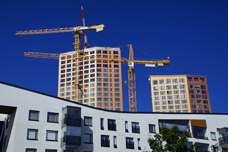 A construction site with two yellow cranes towering over newly constructed or under-construction high-rise buildings. These buildings have numerous windows and a modern design look. The foreground includes a multi-story building with unique geometric windows and a tree with green leaves.