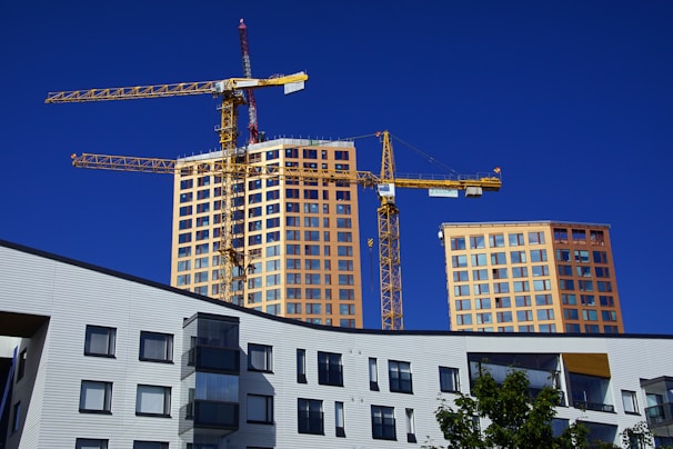 A construction site with two yellow cranes towering over newly constructed or under-construction high-rise buildings. These buildings have numerous windows and a modern design look. The foreground includes a multi-story building with unique geometric windows and a tree with green leaves.