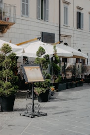 An outdoor restaurant setup with potted plants and white umbrellas lining the sidewalk. The facade of the building features a sign for the restaurant, flanked by light-colored walls and shuttered windows. A menu stand is prominently displayed on the pavement.